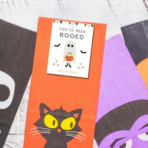 Puede incluir: Tres bolsas de golosinas de Halloween con diferentes dise&ntilde;os. La primera bolsa es naranja con un gato negro y el texto "You've Been Booed". La segunda bolsa es morada con una cara de monstruo blanco y negro. La tercera bolsa es negra con una calavera y huesos blancos.