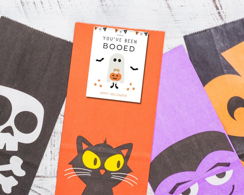 Puede incluir: Tres bolsas de golosinas de Halloween con diferentes dise&ntilde;os. La primera bolsa es naranja con un gato negro y el texto "You've Been Booed". La segunda bolsa es morada con una cara de monstruo blanco y negro. La tercera bolsa es negra con una calavera y huesos blancos.