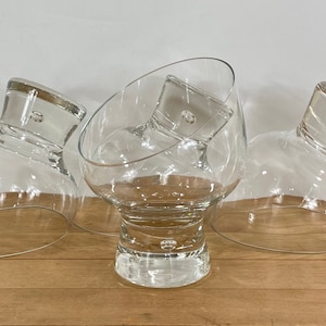 May include: Three clear glass cocktail glasses with a unique design. The glasses have a wide, rounded bowl and a thick, angled stem. They are arranged on a light brown wooden surface against a white background.