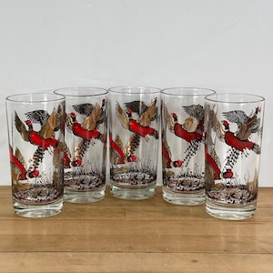 May include: Six clear drinking glasses with a pheasant design. Each glass features a red, gold, and black pheasant in flight, with detailed feathers. The glasses are tall and cylindrical, suitable for water or cocktails.