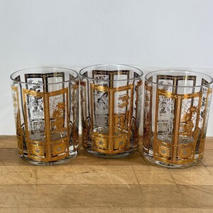 May include: Three clear glass tumblers featuring a repeating gold and black decorative pattern of rectangular frames and intricate floral motifs. Each glass has a cylindrical shape with a flat base and a smooth rim, showcasing a consistent design.