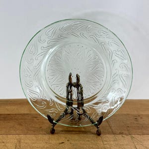 May include: A clear glass plate featuring a central sunburst pattern surrounded by a border of embossed swirling paisley-like motifs. The plate is held upright by a dark metal display stand, showcasing the intricate texture and transparency.