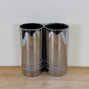 May include: Two chrome-finished cylindrical containers with a shiny, reflective surface. The text "Snap-on" is displayed in a silver, glittery font near the base of each container. The containers are set against a plain white wall and a wooden surface.