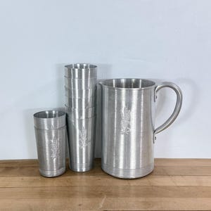 May include: A set of vintage aluminum drinkware, including a pitcher with a handle and several tall glasses. Each piece features an embossed wheat design. The pitcher and glasses are silver-colored and sit on a wooden surface.