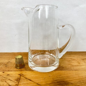 May include: A clear glass pitcher with a handle and a pouring spout. A small, gold-colored thimble sits next to the pitcher on a wooden surface. The pitcher is approximately 8 inches tall.