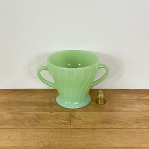 May include: A light green, two-handled sugar bowl with a ribbed design. A small, gold thimble sits to the right of the bowl. The bowl is made of glass and has a rounded base.