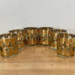 May include: A set of six clear glass tumblers featuring a repeating gold filigree pattern with small green diamond-shaped accents. Each glass has a wide cylindrical shape with a thick base and a gold-rimmed top edge, arranged in a semi-circle.