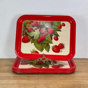 May include: A stack of rectangular metal serving trays with a bright red border and a central illustration of red strawberries with green leaves on a cream background. The top tray is held upright by a decorative gold-toned metal display stand.