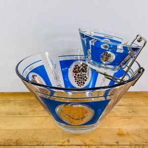 May include: A vintage glass serving bowl set with a blue and gold grape design. The set includes a large bowl and a smaller bowl suspended in a metal holder. The bowls have a clear glass body with blue and gold accents, perfect for serving snacks or dips.