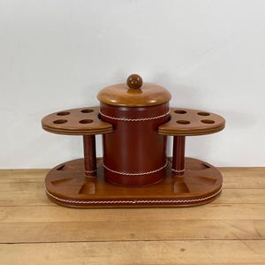 May include: A wooden pipe stand with a central container and two side platforms, each with multiple holes. The stand is made of brown wood with a reddish-brown container and a light-colored lid. White stitching accents the container and base.