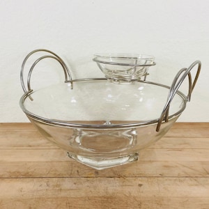 May include: A clear glass serving bowl with a silver metal rim and handles. A smaller glass bowl sits atop the larger bowl, held in place by a silver metal frame. The bowl has a hexagonal base.