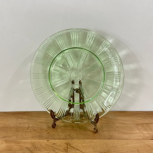 May include: A vintage green glass serving plate with a radial design, resting on a decorative metal stand. The plate features a central circular area and radiating lines, creating a sunburst effect. The plate is displayed on a wooden surface.