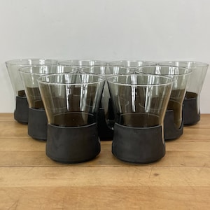 May include: A set of clear drinking glasses with a dark gray base. The glasses have a conical shape and are arranged in a group on a wooden surface. The glasses are empty.