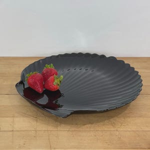 May include: A black, shell-shaped serving dish with a scalloped edge. Three fresh strawberries with green stems rest on the dish. The dish is on a wooden surface.