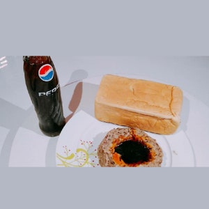Può includere: Una bottiglia di vetro di Pepsi con un logo rosso, bianco e blu, una pagnotta di pane e una polpetta con salsa scura su un piatto bianco. Il cibo è disposto su una superficie bianca.
