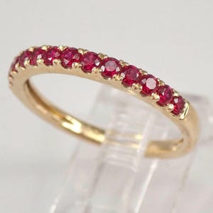 May include: A gold ring featuring a line of small, round, bright red gemstones. The ring is presented on a clear stand, highlighting the polished gold and the vivid colour of the gemstones.