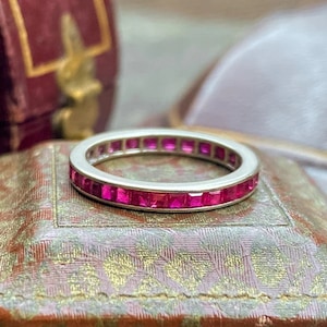 May include: A silver eternity band ring with a row of square-shaped pink ruby gemstones.