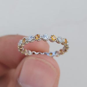 Op de afbeelding: Een zilverkleurige ring met een afwisselend patroon van heldere witte stenen en geel-oranje citrienstenen in een klauwzetting. De tekst Rings Of Memory is in kleine zwarte letters gedrukt in het midden van de ringband.