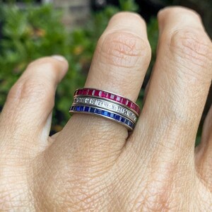 May include: Three eternity bands with red, white, and blue gemstones. The bands are stacked on a finger.