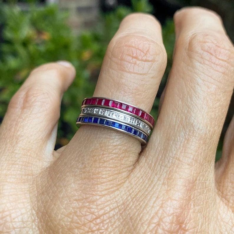 May include: Three eternity bands with red, white, and blue gemstones. The bands are stacked on a finger.