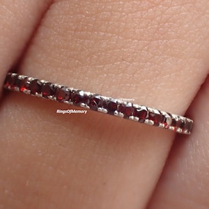 Peut inclure: Une bague en argent ornée d'une rangée continue de petites pierres précieuses rondes, rouge foncé. Les pierres sont rapprochées, créant une ligne de couleur autour de la bague. La bague est portée sur un doigt.