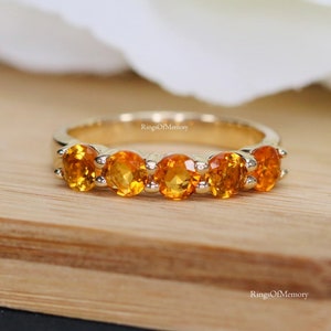 Puede incluir: Un anillo de banda de oro con cinco piedras preciosas naranjas. El anillo está colocado sobre una superficie de madera.