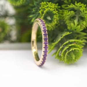 Peut inclure: Une bague en or ornée de petites pierres précieuses rondes et violettes. La bague est mise au point sur un fond flou de feuillage vert et une surface blanche. Les pierres sont disposées en cercle.