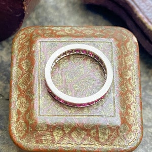 May include: A close-up of a pink gemstone eternity band ring set in white metal. The ring is sitting on a brown patterned surface.