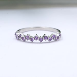 May include: A silver ring featuring a row of small, round, purple gemstones. The gemstones are evenly spaced along the band, creating a sparkling, elegant design. The ring is set against a plain white background.