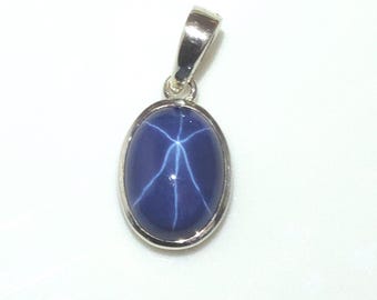 May include: A blue star sapphire pendant with a silver bail. The oval-shaped gemstone displays a six-rayed star pattern. The pendant is set in a silver bezel.