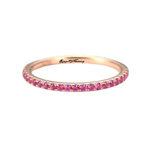 May include: Rose gold band ring set with a continuous row of small, bright pink gemstones. The ring has a smooth, polished finish and a delicate design. The text "Ginger Of Memory" is visible on the ring.