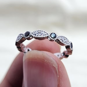 Op de afbeelding: Een zilveren ring met een herhalend patroon van kleine, heldere en zwarte stenen. De ring wordt door een vinger omhoog gehouden, waarbij de nadruk ligt op het ingewikkelde ontwerp en het contrast tussen het zilver en de stenen.