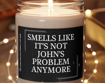 May include: A brown soy candle with a black label that reads "Smells like it's not John's problem anymore". The candle is lit and sitting on a wooden coaster.