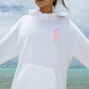 K&ouml;nnte beinhalten: Wei&szlig;er Oversize-Hoodie mit einem pinkfarbenen Blitz-Motiv auf der Vorderseite. Der Hoodie hat eine K&auml;ngurutasche und eine Kapuze mit Kordelzug.