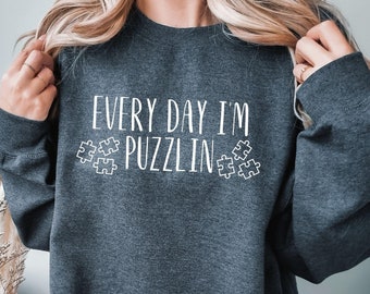 May include: Dark gray sweatshirt with the text "EVERY DAY I'M PUZZLIN" in white, along with puzzle piece graphics. The sweatshirt is a crew neck style and appears to be made of a soft material.