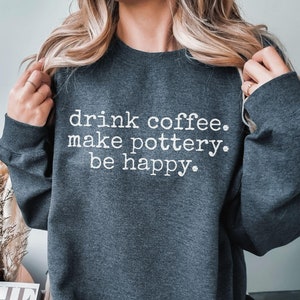 May include: A dark gray sweatshirt with the text "drink coffee. make pottery. be happy." printed in white.