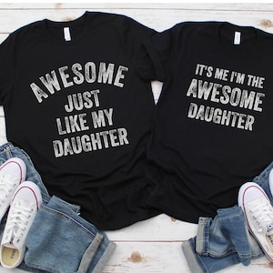 May include: Two black t-shirts with white text. The first shirt says "Awesome Just Like My Daughter". The second shirt says "It's Me I'm The Awesome Daughter".