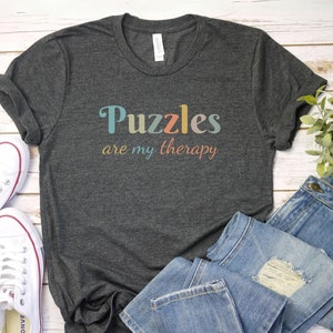 May include: A dark gray t-shirt with the text "Puzzles are my therapy" in colorful letters. The shirt is laid out on a white wooden surface with a pair of white sneakers and blue jeans.