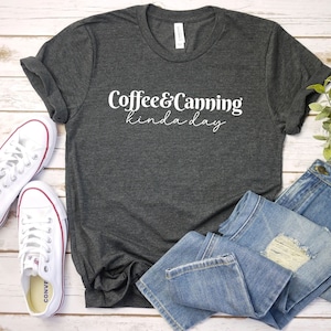 Op de afbeelding: Een donkergrijs T-shirt met de tekst "Coffee & Canning kinda day" in witte letters. Het shirt ligt op een wit houten oppervlak met een paar witte sneakers en een blauwe jeans.