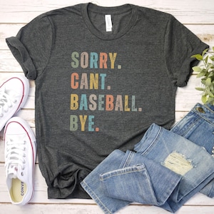May include: A dark gray t-shirt with the text "SORRY. CANT. BASEBALL. BYE." in colorful retro-style lettering.