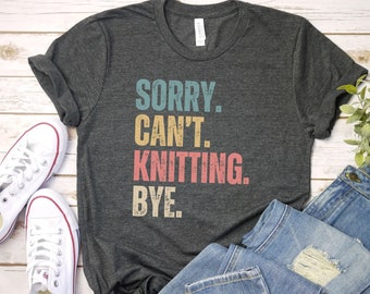 May include: A dark gray t-shirt with the text "SORRY. CAN'T. KNITTING. BYE." in a vintage style font with colorful accents.