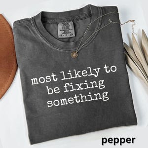 May include: A charcoal gray short-sleeve t-shirt with a crew neckline. The shirt features the white text "most likely to be fixing something" printed on the front. A Comfort Colors brand label is visible inside the collar, and a thin necklace rests on the fabric.