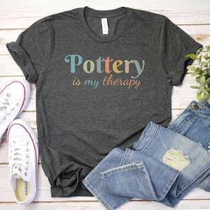 May include: A dark gray t-shirt with the text "Pottery is my therapy" in colorful letters. The shirt is laid out on a white wooden surface with a pair of white sneakers and blue jeans.