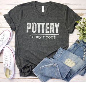 May include: A dark gray t-shirt with the text "POTTERY is my sport" in white lettering. The shirt is laid out on a white wooden surface with a pair of white sneakers and blue jeans.