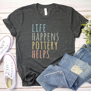 May include: A dark gray t-shirt with the text "Life Happens Pottery Helps" in rainbow colors. The shirt is folded and displayed on a white wooden surface with a pair of white sneakers and blue jeans.