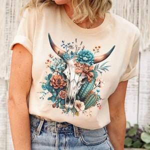 Puede incluir: Una camiseta beige con un dise&ntilde;o floral que presenta una calavera con cuernos y flores turquesas y naranjas.