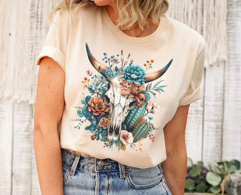 Puede incluir: Una camiseta beige con un dise&ntilde;o floral que presenta una calavera con cuernos y flores turquesas y naranjas.