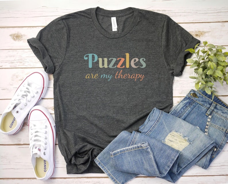 May include: A dark gray t-shirt with the text "Puzzles are my therapy" in colorful letters. The shirt is laid out on a white wooden surface with a pair of white sneakers and blue jeans.