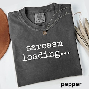 May include: A dark gray t-shirt with the words "sarcasm loading..." printed in white. The shirt has a crew neck and short sleeves. A silver necklace with a pendant is draped across the shirt. The word "pepper" is printed at the bottom.