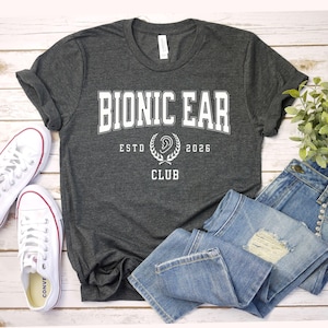 May include: Dark gray t-shirt with the text "BIONIC EAR CLUB" in white, along with the year "2026" and a graphic of an ear. The shirt is paired with blue jeans and white sneakers.